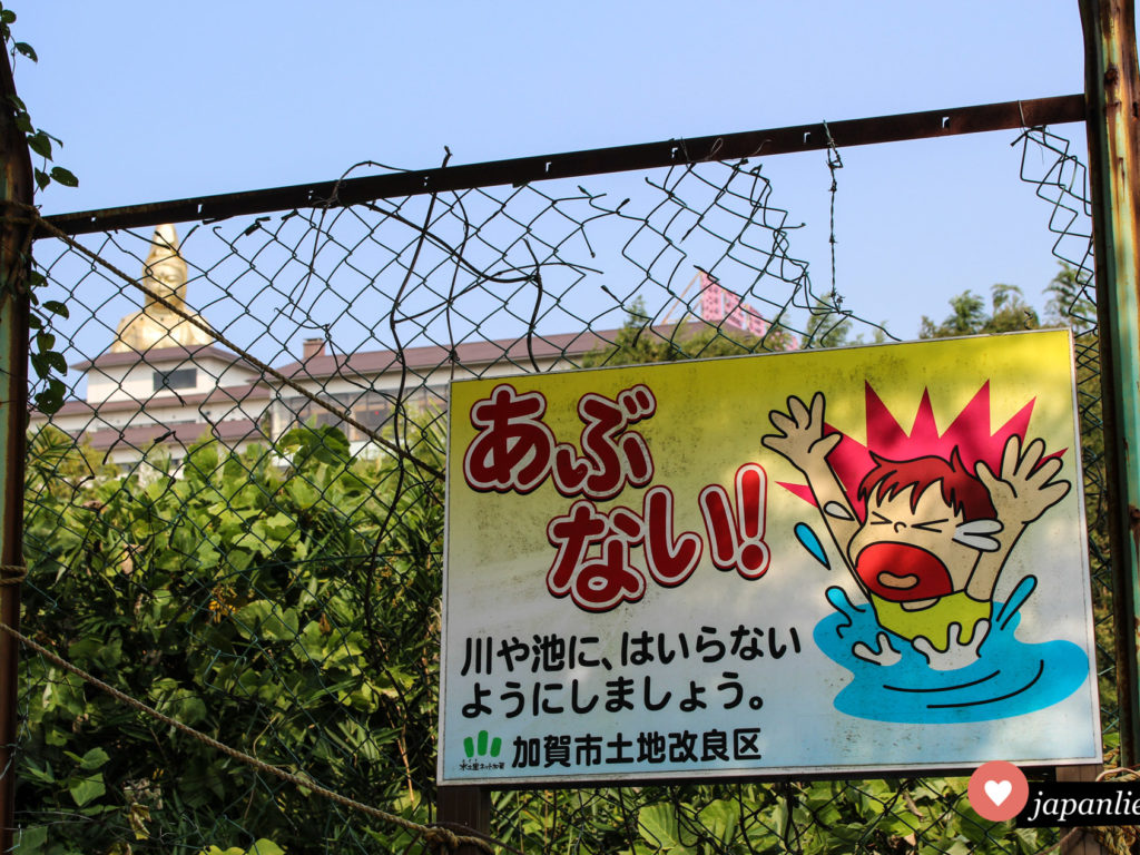 Ein Schild in Kaga-Onsen warnt davor, dass Schwimmen an dieser Stelle zu gefährlich ist. Im Hintergrund wacht eine riesige, goldene Buddhastatue darüber, ob dieses Verbot auch eingehalten wird.