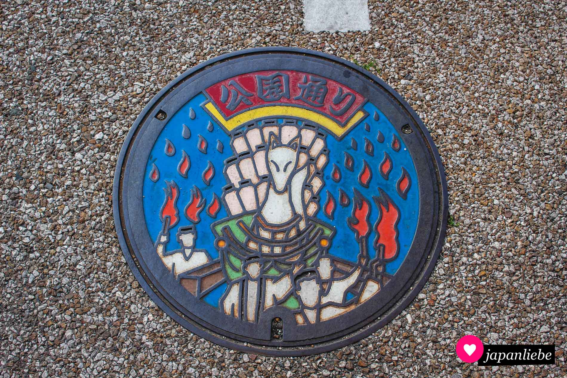 Eine o-mikoshi-Göttersänfte mit Fuchs und Laternen wird bei diesem Kanaldeckelbild durch die Straßen getragen.